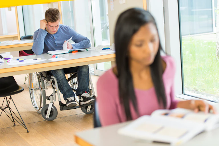 a young white man in a wheelchair sits at a table reading behind a woman sitting and reading