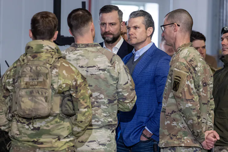 Two men in suits talk with three men in army camouflage uniforms.