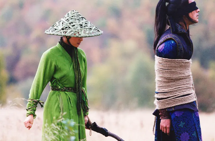 Una mujer con atuendo tradicional chino y una daga sigue a un hombre atado y con los ojos vendados.