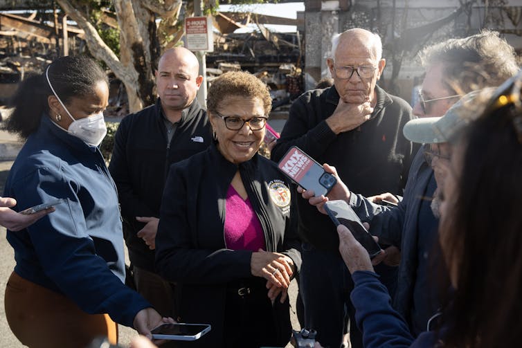 A woman speaks into microphones, with burned homes in the background.