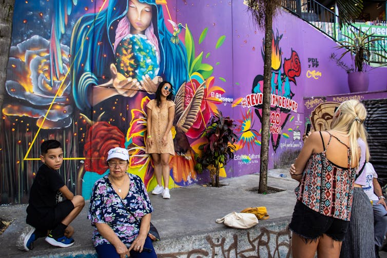 A tourist poses for a picture next to a multicoloured wall.