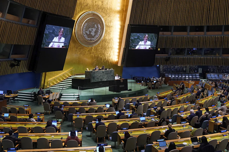 A woman in a white jacket addresses the United Nations and is seen on jumbo screens behind her to the left and right.