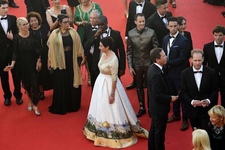 Woman wearing dress with a depiction of a city skyline walking among a crowd of people on a red carpet.