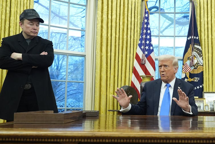 A white man with orange-tinged skin and white hair gestures while speaking from behind a large wooden desk. Another white man in a black ball cap and coat stands with his arms crossed beside him