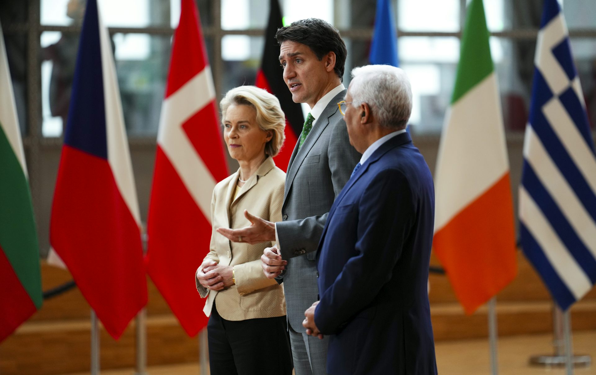 A white man with dark hair, flanked by a white woman with blonde hair and a man with brown skin and white hair, speaks to someone off-camera while standing in front of a row of flags