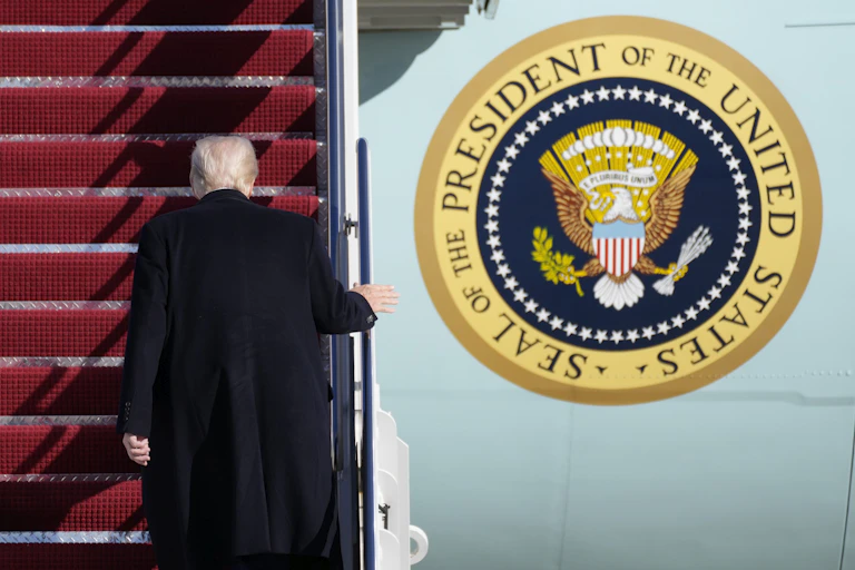 A man with white hair and black coat is seen boarding a plane from behind. The seal of the U.S. president is visible on the side of the plane