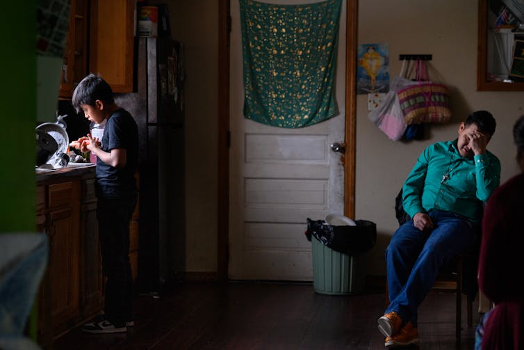 A man sits and covers his face with his hand, while a young boy washes dishes nearby him.