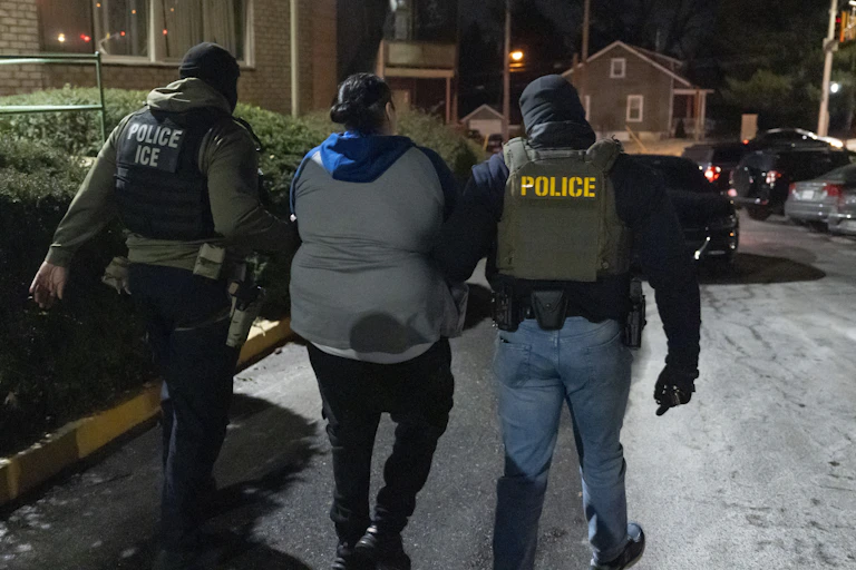 Two men wearing long sleeves and a vest that says 'police' and 'police ICE' on the back walk holding the arms of a person. They are seen from behind.