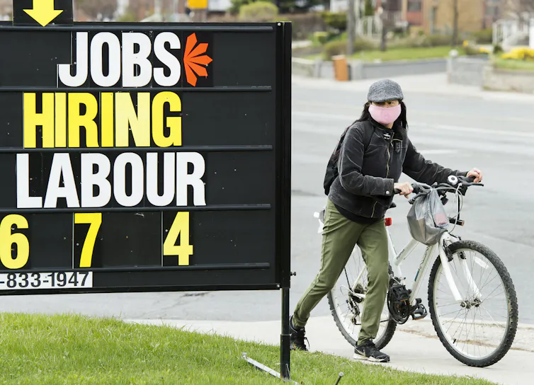 A woman walks a bicycle past a sign that says 'Jobs hiring labour'