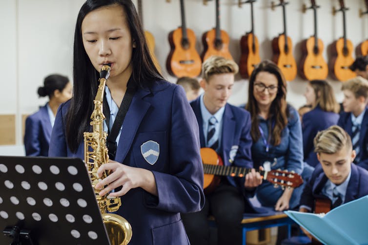 A student plays the saxophone. Other students play instruments in the background. A line of guitars hang on a wall.