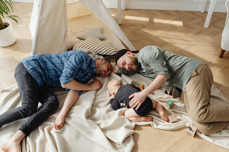 Two men lying on the floor, looking tired, with a baby in a black onesie between them.