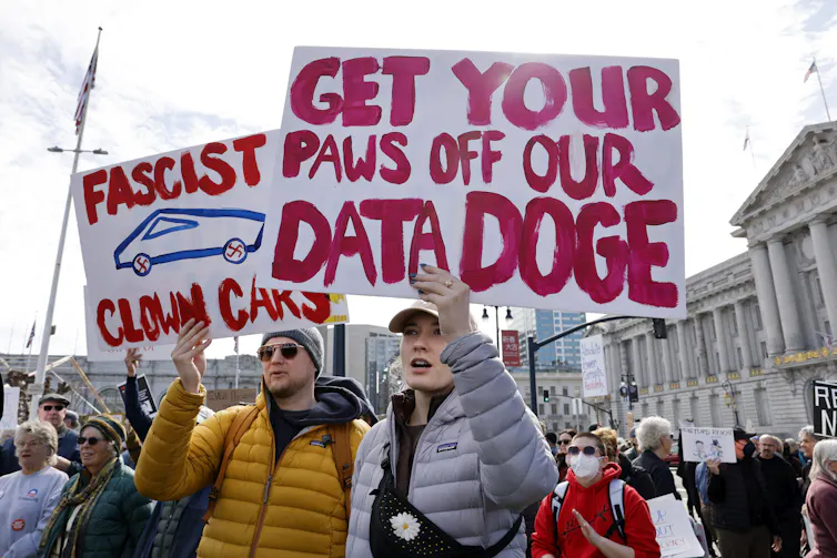Photo of protesters, one holding a placard reading 'GET YOUR PAWS OFF OUR DATA DOGE'.