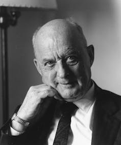 A black-and-white portrait of a bald man in a jacket and tie, posing with his fist against his chin.