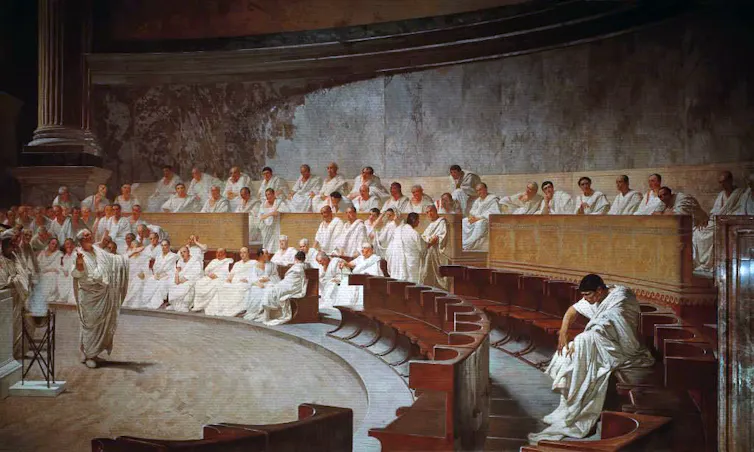 Un hombre vestido con una toga blanca habla ante otros en el Senado romano.