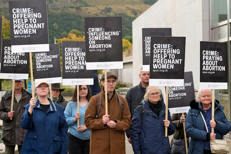 A group of people holding printed signs that list the various so-called crimes they believe fall under the safe access zones law.