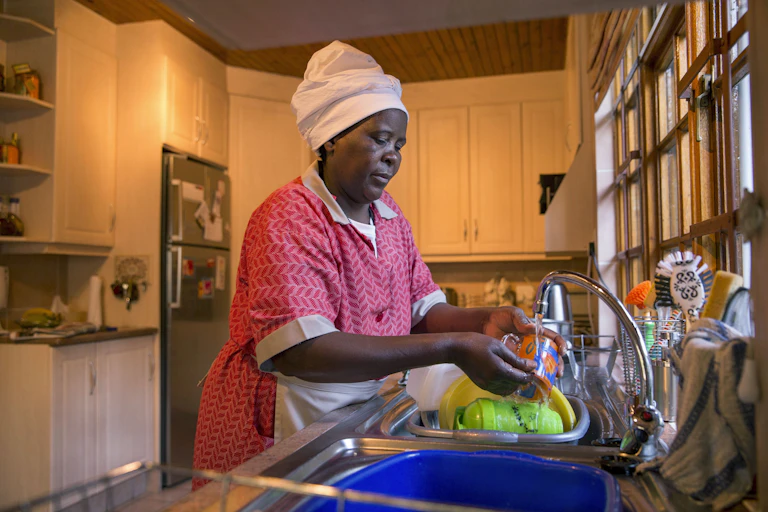 A woman wearing a headscarf, overall and apron, washing items at a sink in a kitchen