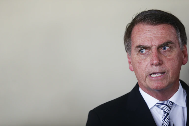 Jair Bolsonaro standing against a white wall.
