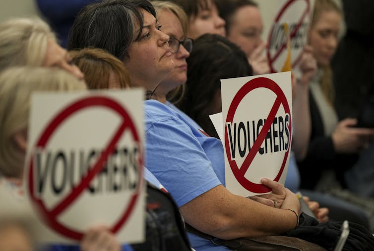 People sit holding signs with the word 'voucher' crossed out by a red line while staring ahead