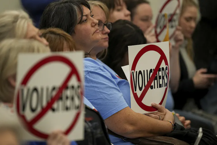 People sit holding signs with the word 'voucher' crossed out by a red line while staring ahead