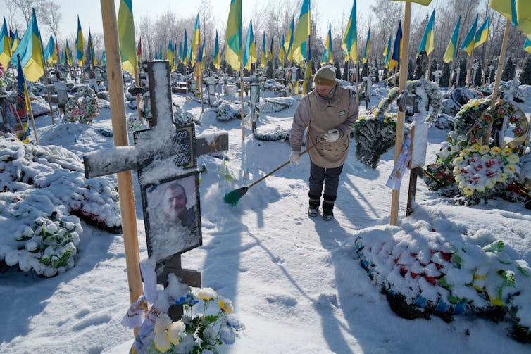 A person brushes snow at a gravesite that has photos of people on crosses and blue and yellow flags.