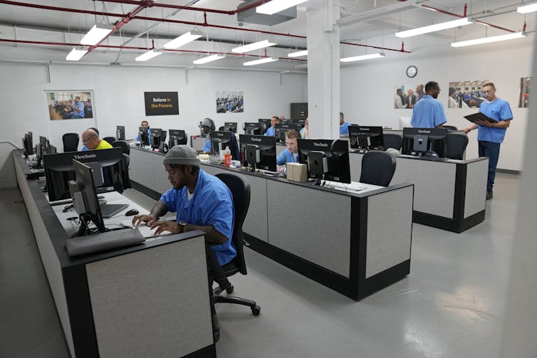 People in prison clothes sit at desks with computers and monitors.