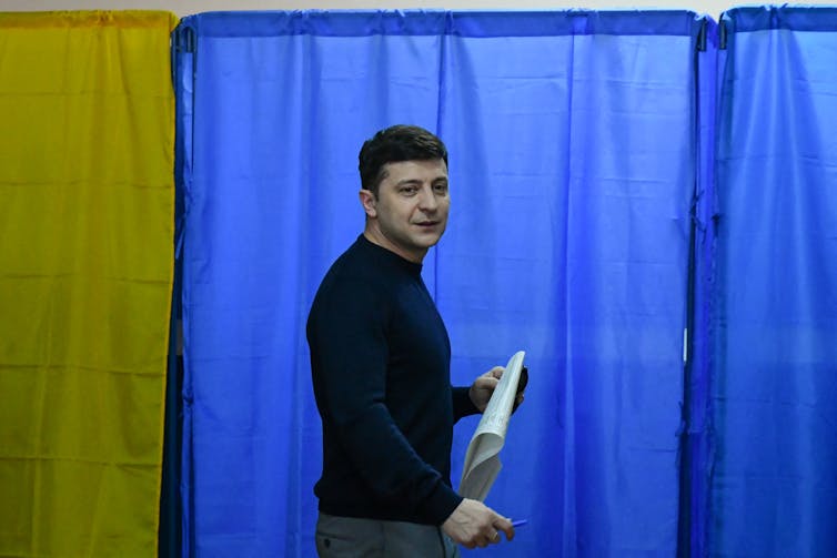 A man holds a piece of paper in front of a yellow and blue curtain.