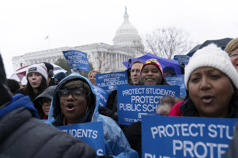 People hold signs that say 'protect students protect public schools' in front of a domed building.