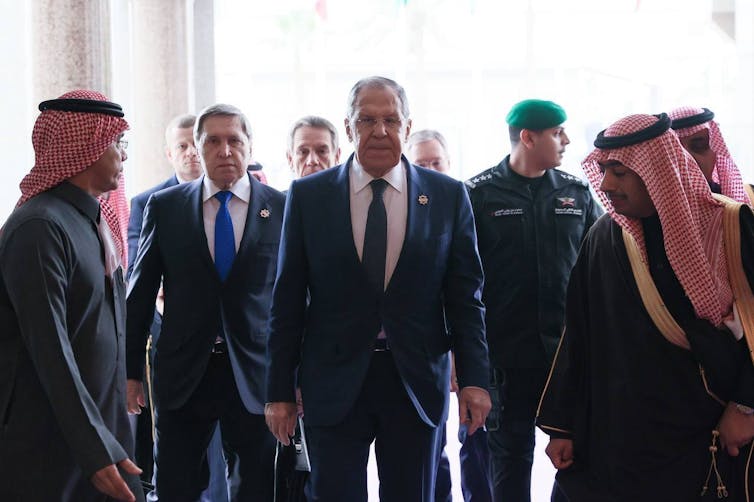 A man in a suit walks toward the camera flanked by men in traditional Arab Gulf headscarves.