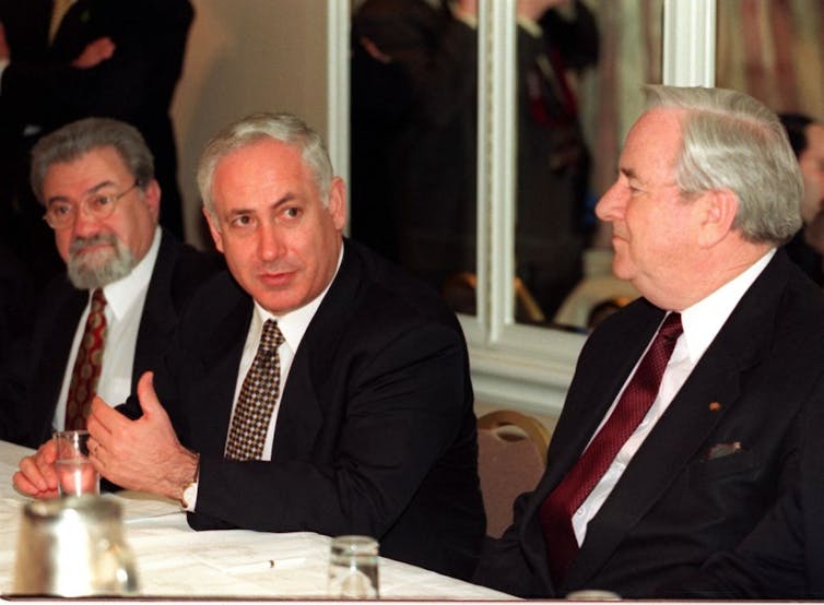 Three men in suits and ties sit at a table as the man in the center speaks.