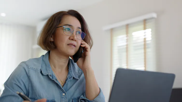 A woman holds a phone to her ear. She sits in front of a laptop.
