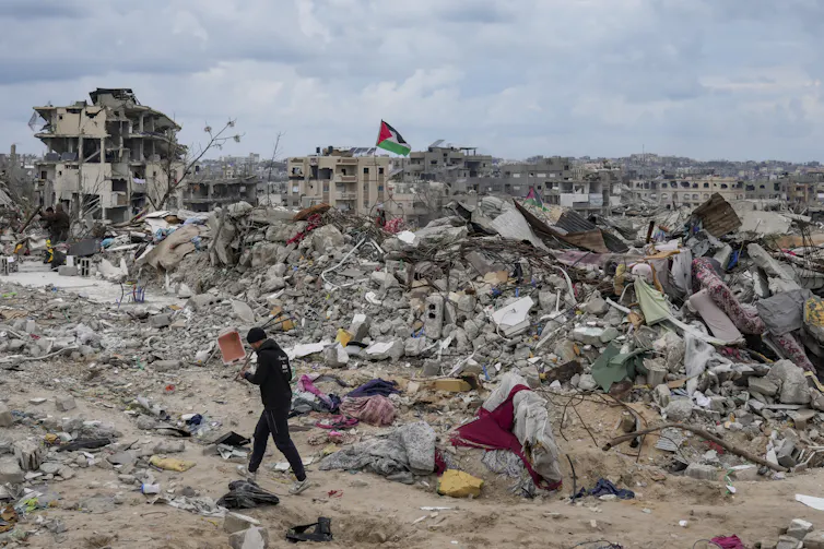 A man walks past rubble of destroyed buildings, a palestinian flag is seen on a pole above the rubble.
