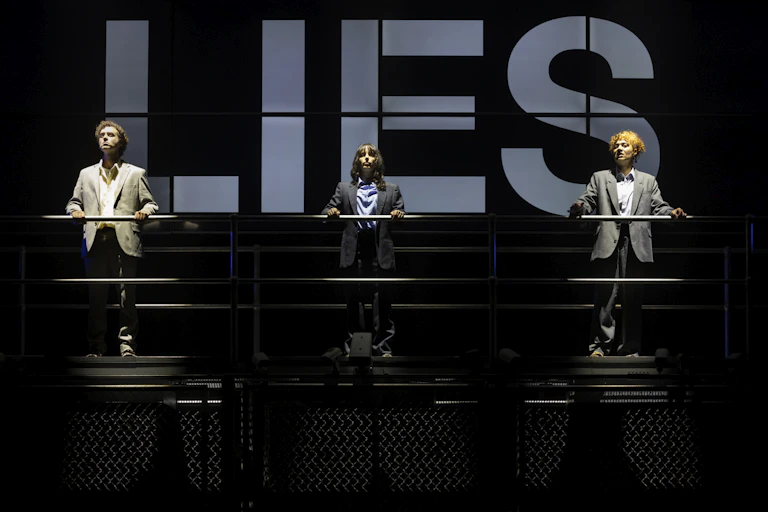 Three people on stage. Behind them reads the word Lies.
