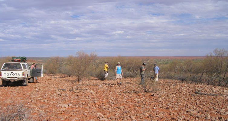 A group of people standing on a red rocky outcrop looking across a flat horizon.