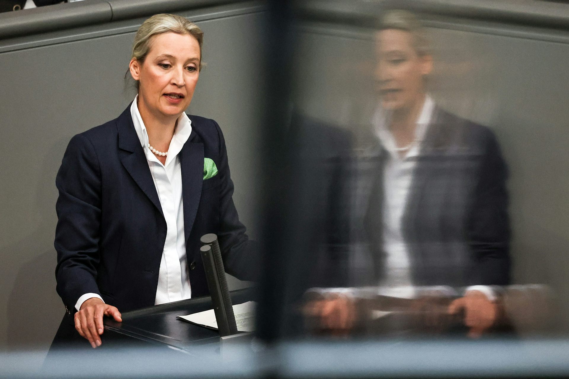 On the left side of the photo, Alice Weidel speaking at a podium, on the right side a slightly distorted reflection of Weidel in glass