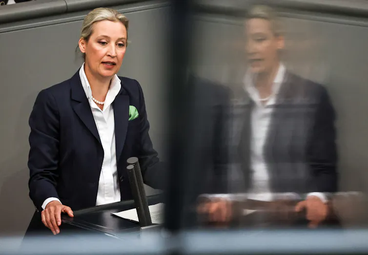 On the left side of the photo, Alice Weidel speaking at a podium, on the right side a slightly distorted reflection of Weidel in glass