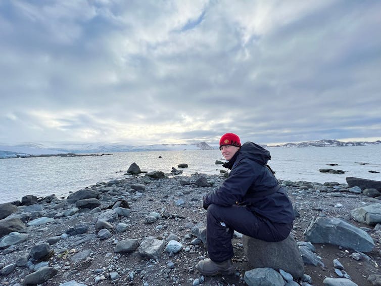 A man wearing dark cold-weather clothing and a bright red hat sits on a rock near water and under a cloudy sky in Antarctica.