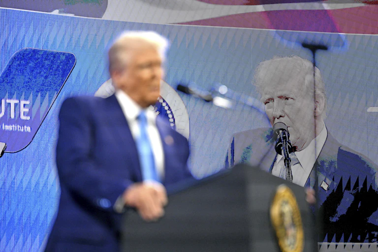 A man with white-ish blond hair is blurred speaking at a podium and in focus on a screen behind him.
