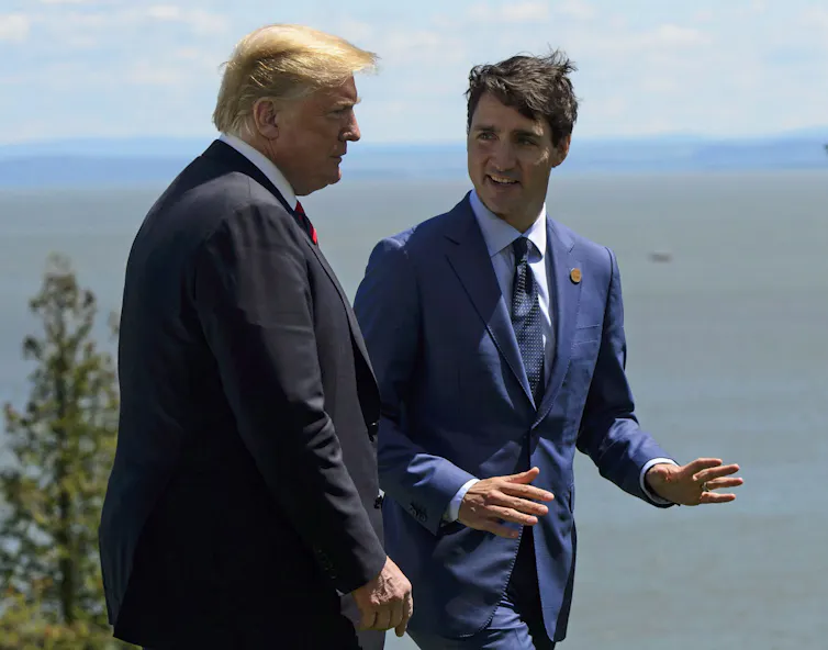 A rotund man with fluffy orange-tinged hair walks next to a slim dark-haired man with a lake in the background.