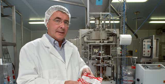 A man in a lab coat and a hair net holds three red and white packets in a small factory.