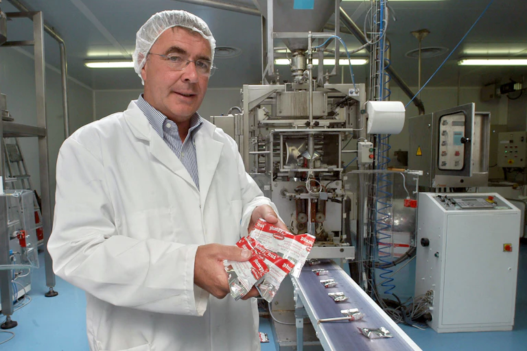 A man in a lab coat and a hair net holds three red and white packets in a small factory.