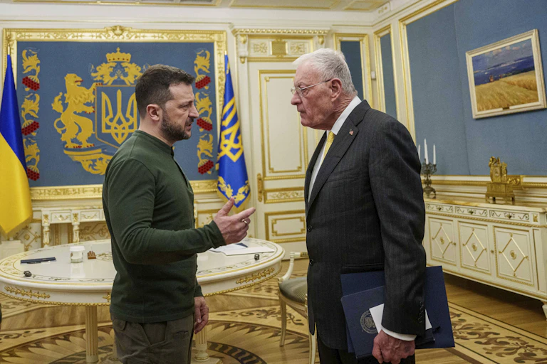 A bearded dark-haired man in a dark green shit speaks to an older tall man in a suit in an ornate office with a blue and yellow flag behind them.