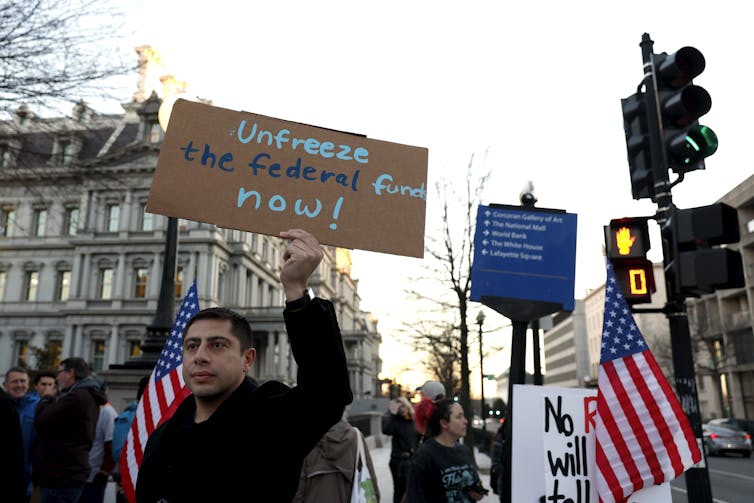 Man in a crowd holds a sign reading 'unfreeze the federal funds now'