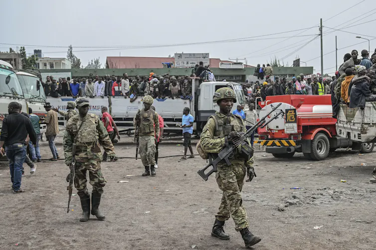 armed soldiers in the foreground, lorries loaded with people in the background on a street