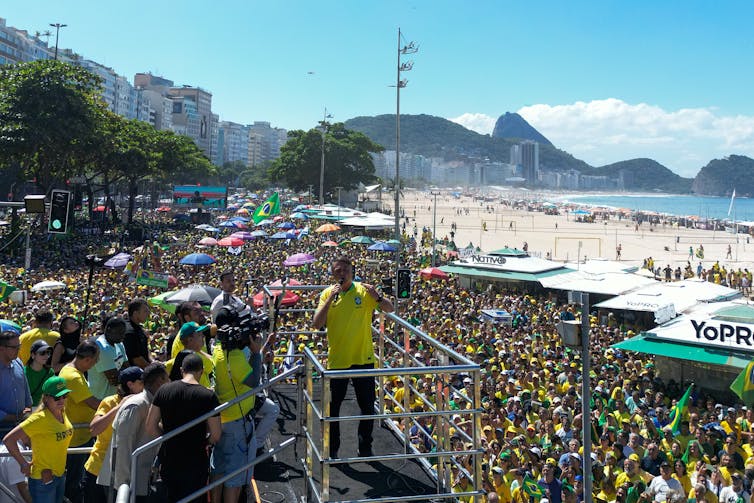 Man in yellow t-shirt speaks to throngs assembled on a beach in Rio de Janeiro.