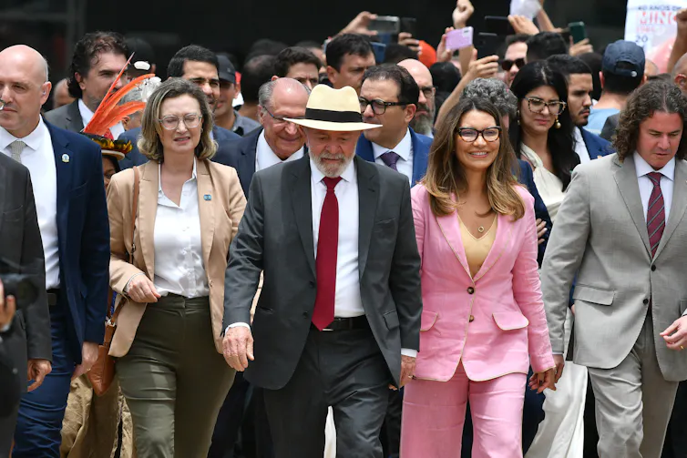 Man in straw hat, accompanied by a woman in a pink suit, as they lead a crowd of people in business attire.