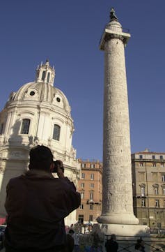A person seen from the back takes a photo of a tall monument with a statue on top, and a domed building in the background.