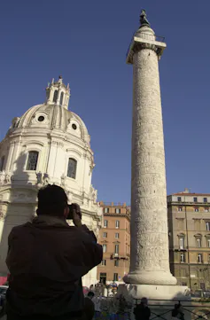 A person seen from the back takes a photo of a tall monument with a statue on top, and a domed building in the background.