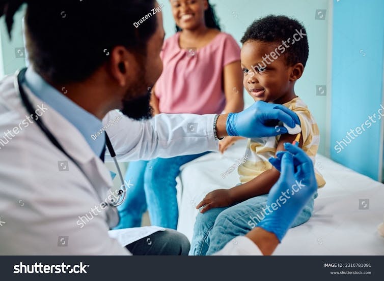 A health-care professional in a white coat and blue gloves give a shot to a small boy, with a woman in the background
