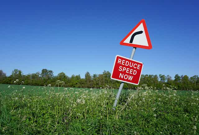 Red and white road sign in the countryside instructing people to slow down.