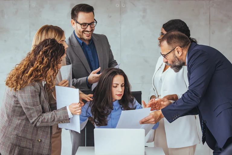 Five adults in business attire are surrounding a colleague who is seated and reading a document.
